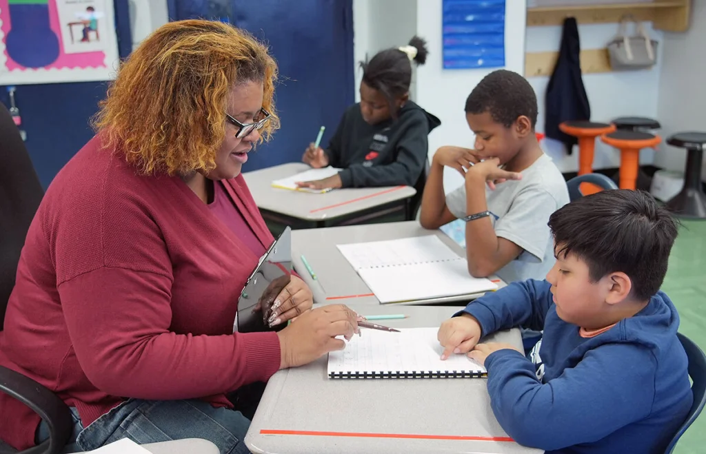 Teacher reviewing student's dictation work while other students work in the background.
