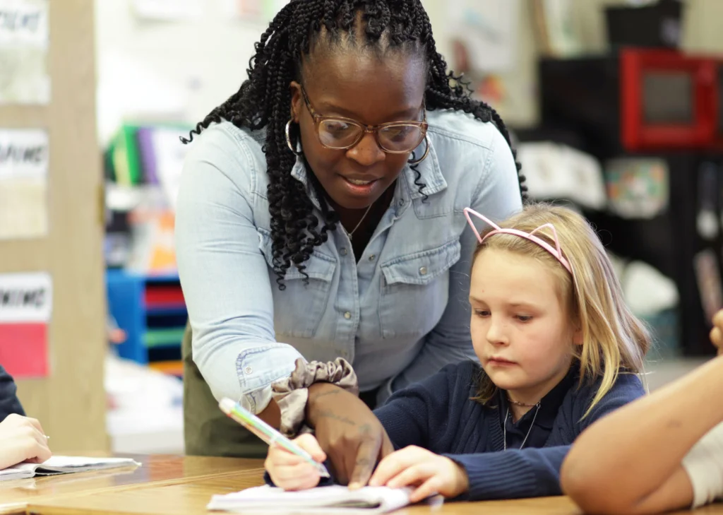 Teacher helping a young student with her worksheet at the student's desk.
